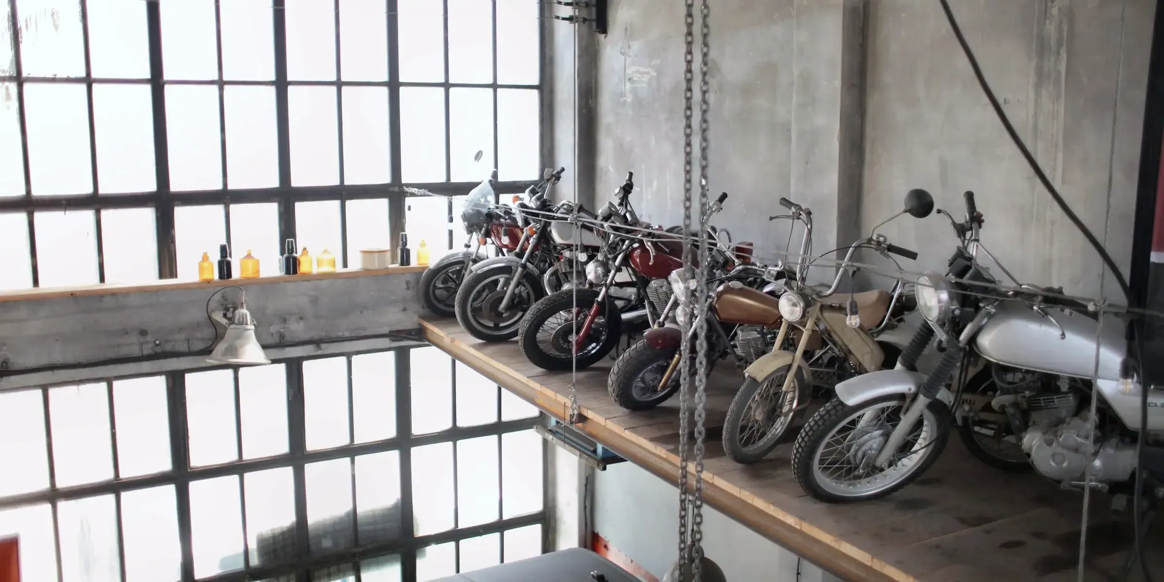 Un atelier à verrière d’usine : une rangée de motos anciennes sur la mezzanine, une chaîne de palan au premier plan