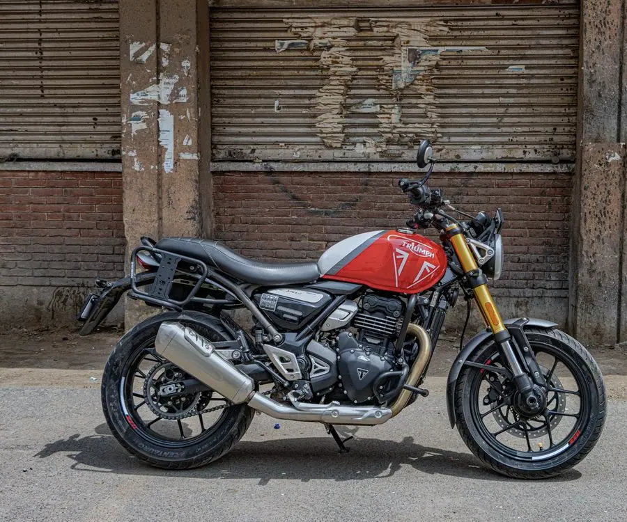 Triumph Scrambler 400 X rouge et noire de profil, fourche dorée et porte-bagages, devant un rideau de fer