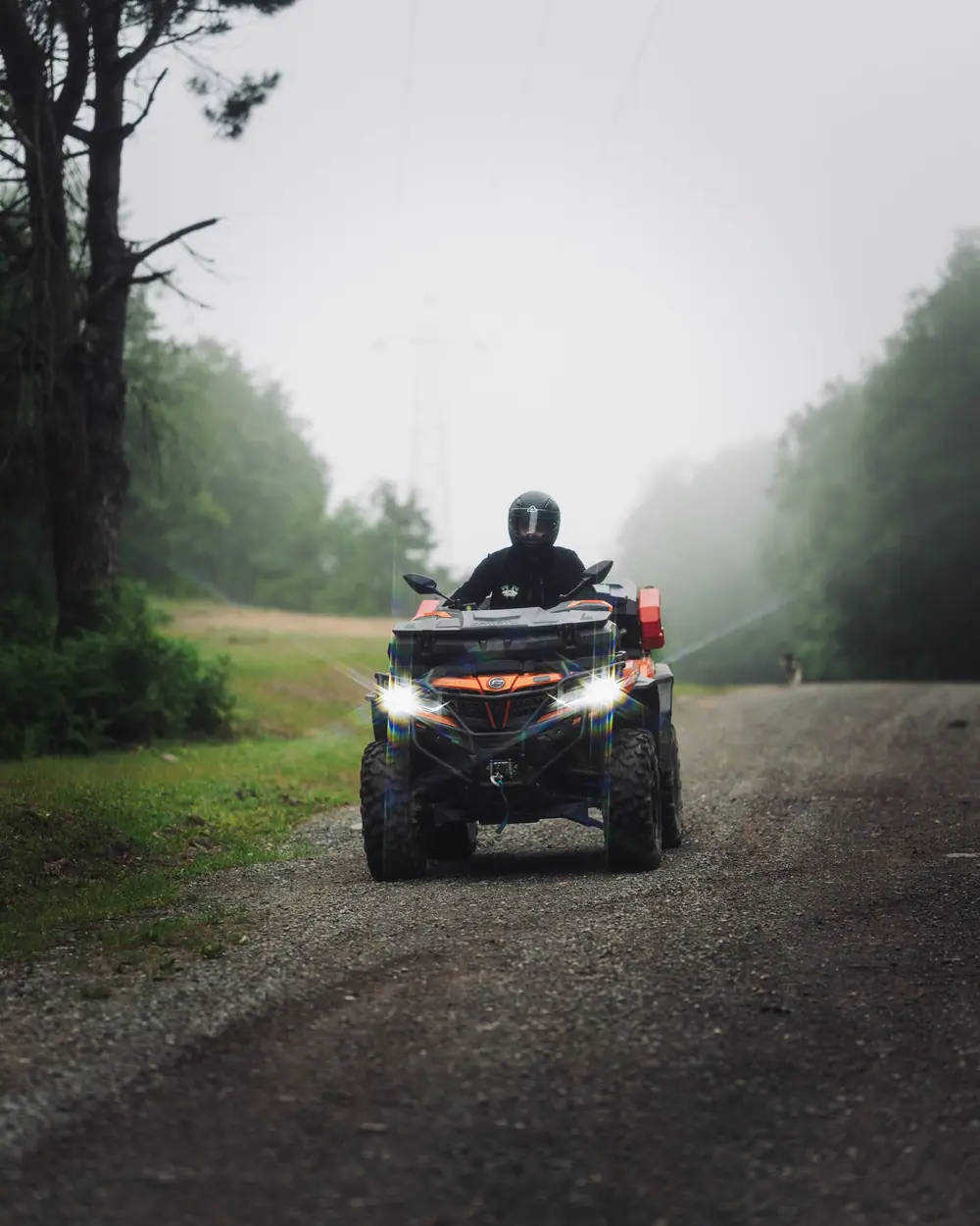 Quad orange phares allumés sur un chemin de forêt dans la brume, pilote casqué