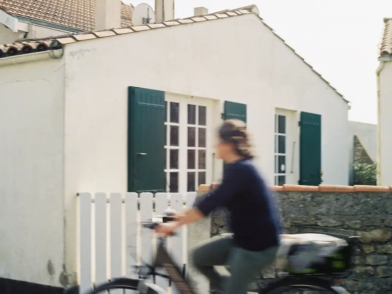 Une cycliste passe devant une maison blanche aux volets verts.