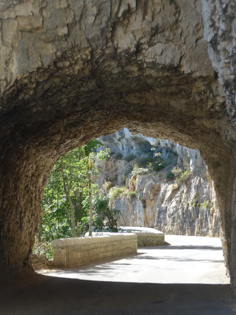 Arche d’un tunnel taillé dans la roche : la route, son parapet, et la falaise d’en face en plein soleil